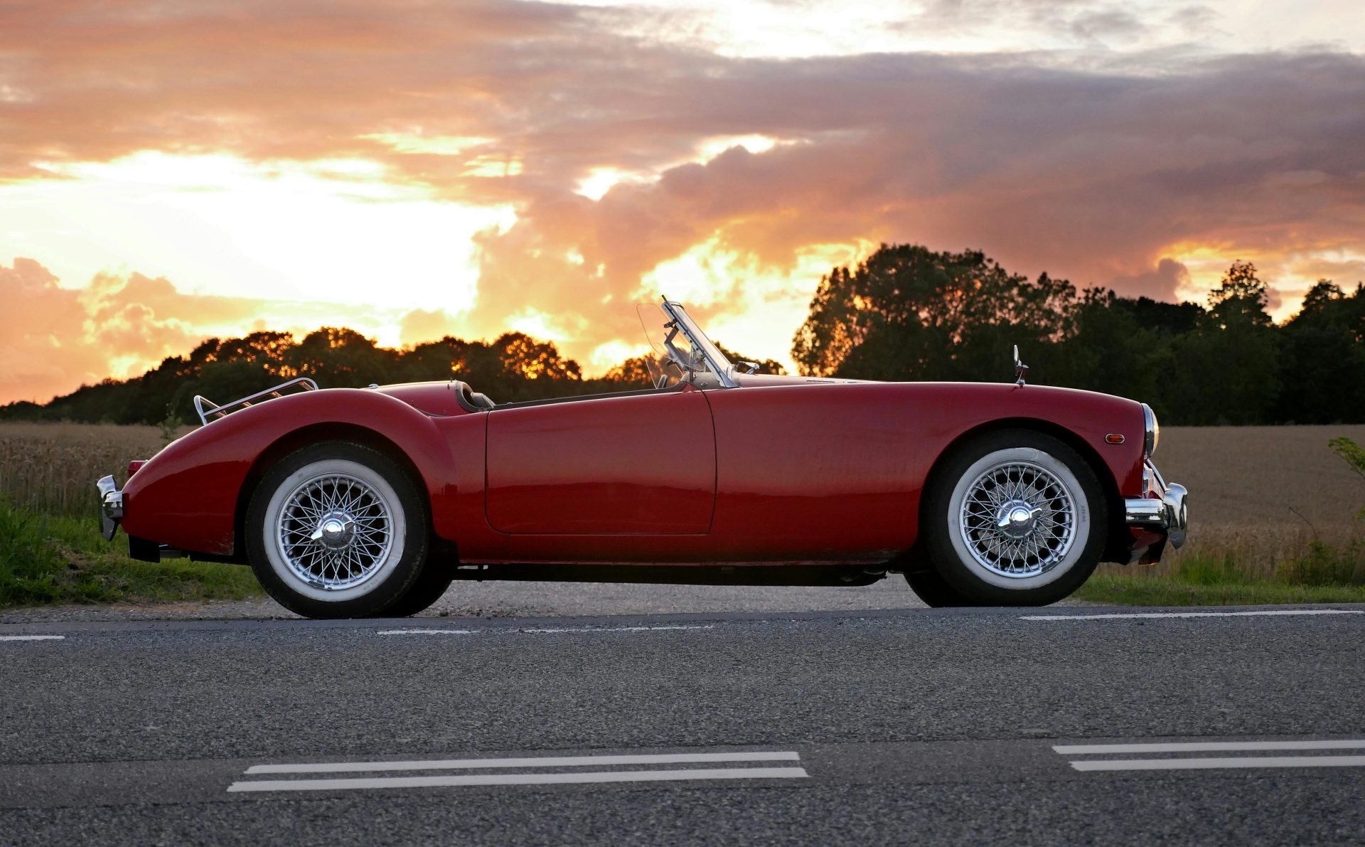 Vintage red convertible car parked on a country road at sunset with dramatic skies.