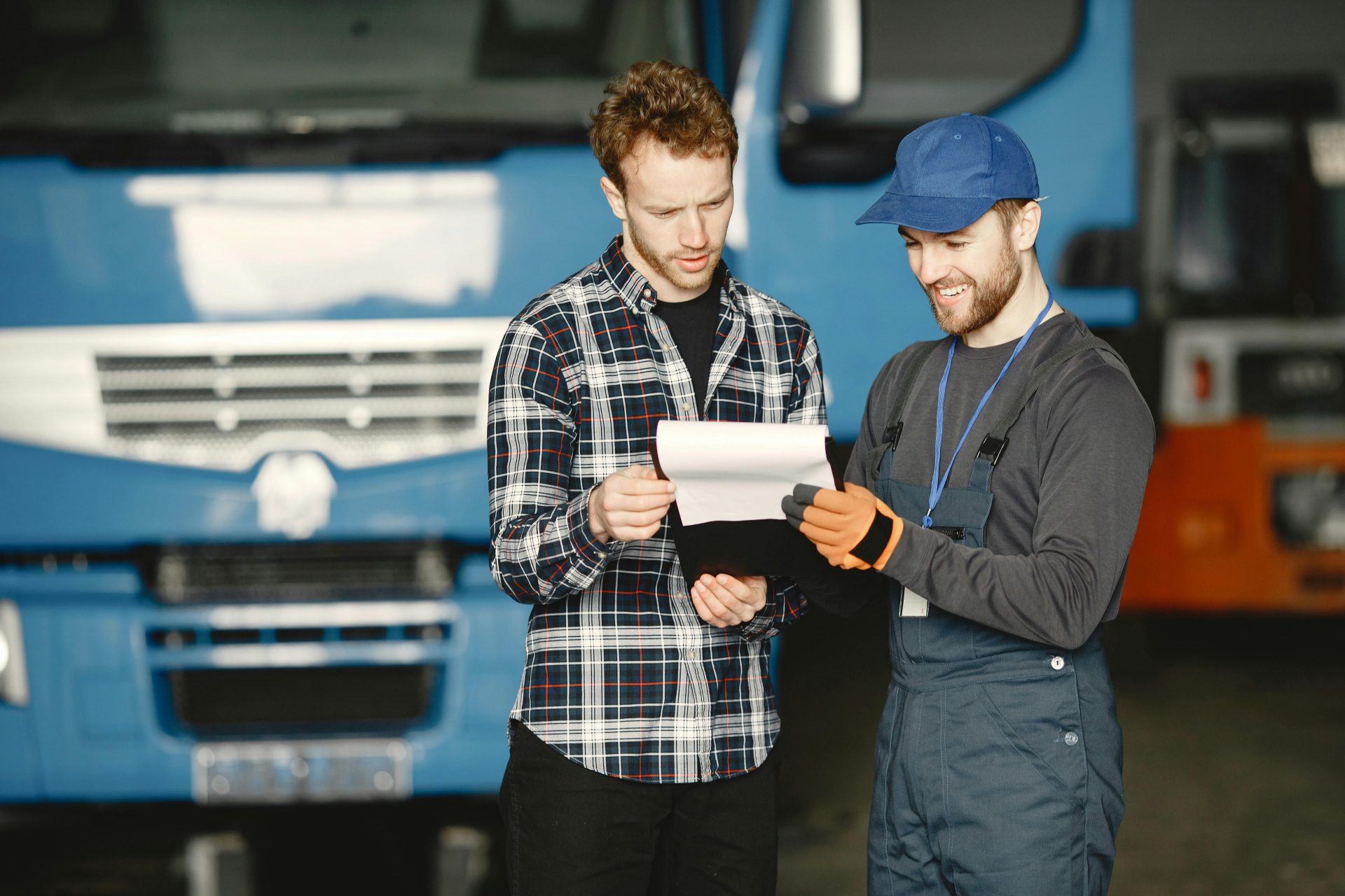 Two men in discussion over vehicle maintenance documents in a garage setting with trucks in the background.