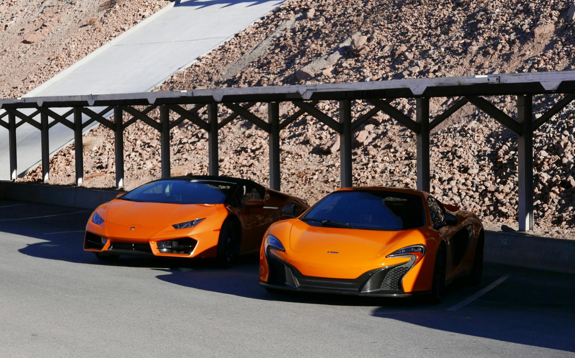 Two orange luxury sports cars parked under a wooden structure in Boulder City, Nevada.